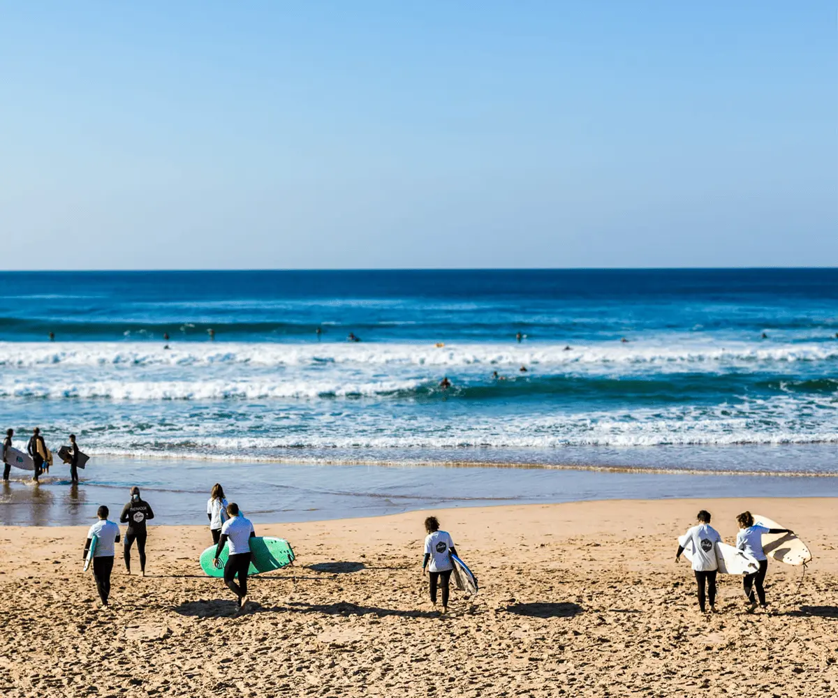 Grupo de alunos da Gecko Surf School a caminhar na praia da Costa da Caparica com pranchas de surf durante uma aula de surf em grupo