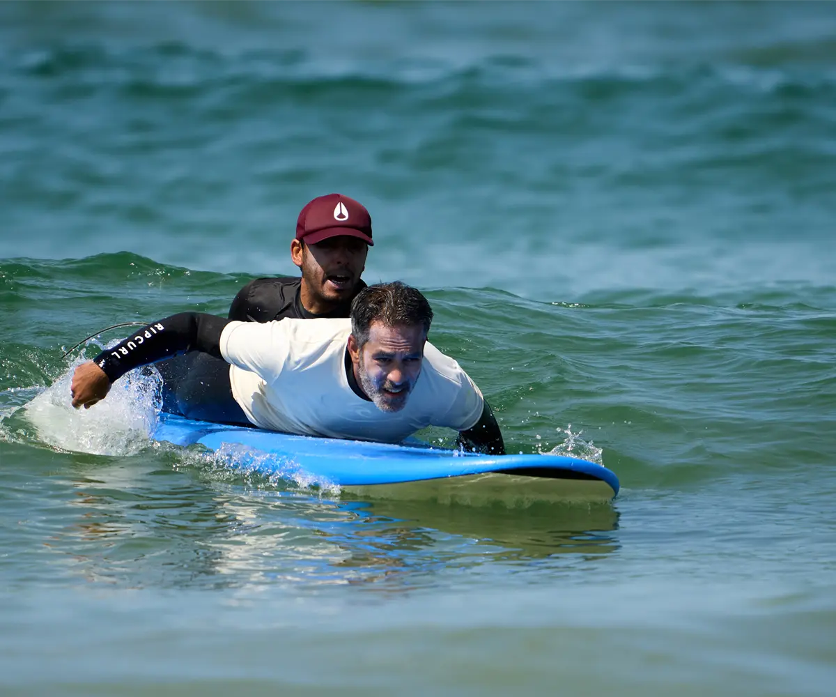 Instrutor da Gecko Surf School a dar uma aula de surf privada na Costa da Caparica, auxiliando um aluno adulto na prancha dentro de água