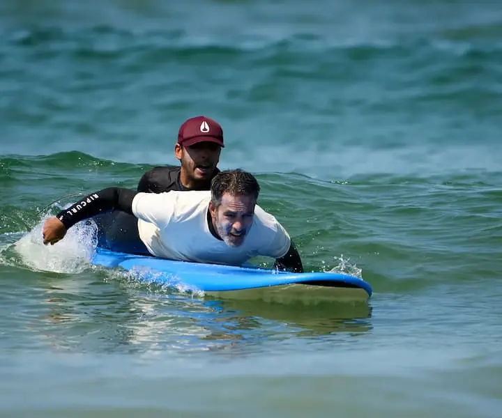Instrutor da Gecko Surf School a dar uma aula de surf privada na Costa da Caparica, auxiliando um aluno adulto na prancha dentro de água